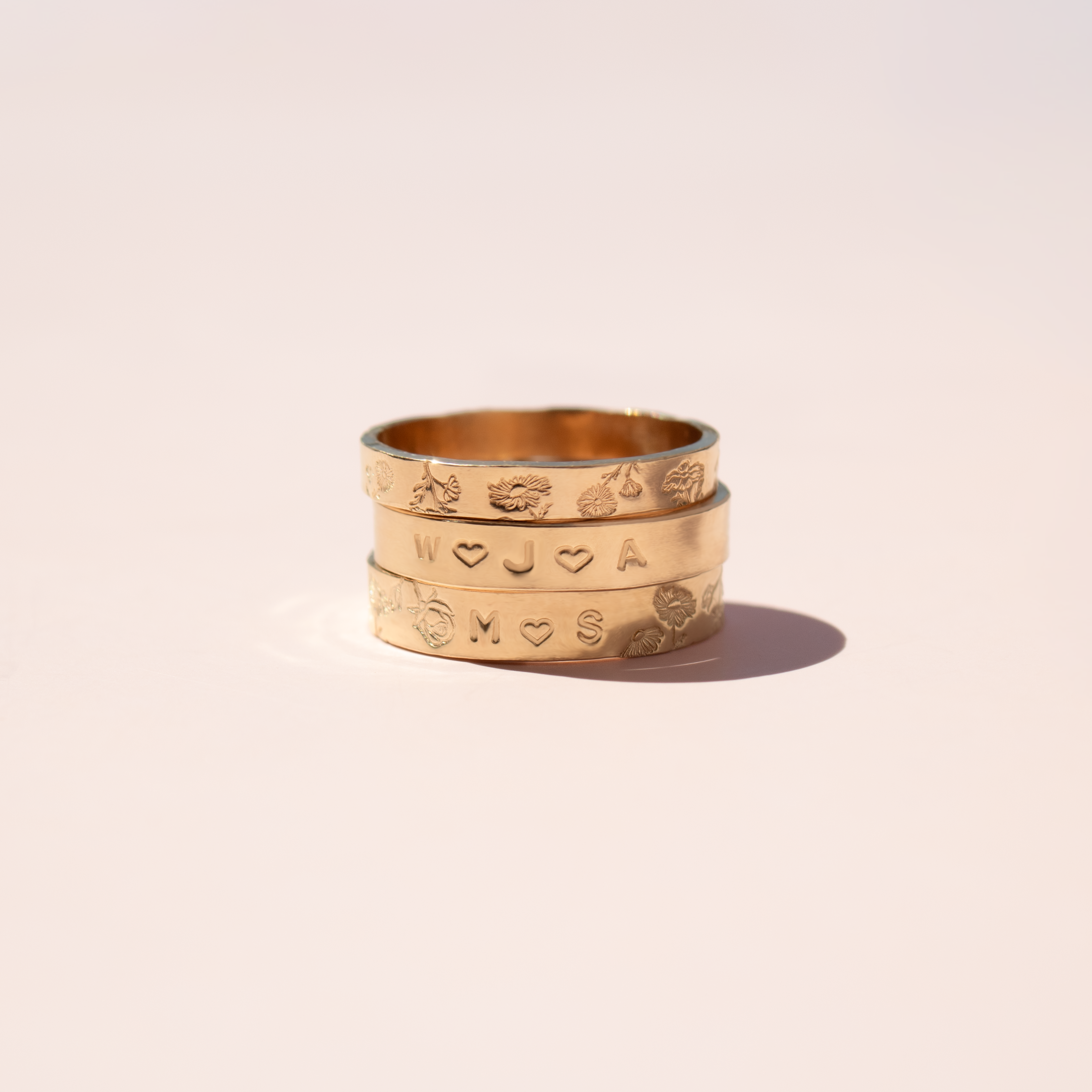 A stack of three gold rings with engraved initials and small heart motifs, displayed on a clean, solid light beige background.